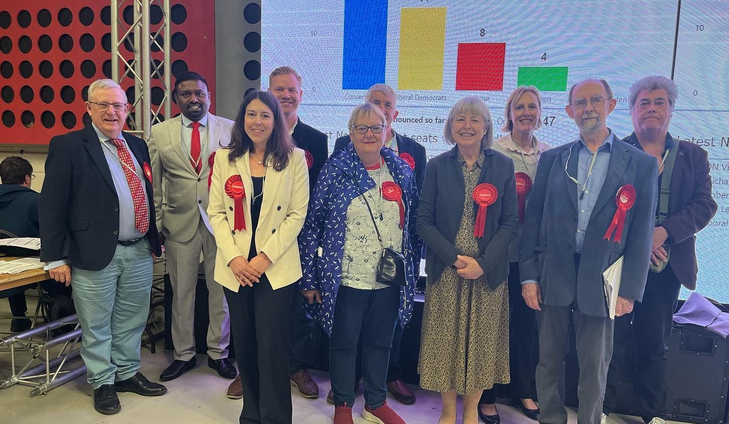 10 people standing in front of a screen which shows coloured bar charts displaying the number of councillors in Broadland, won by each party.